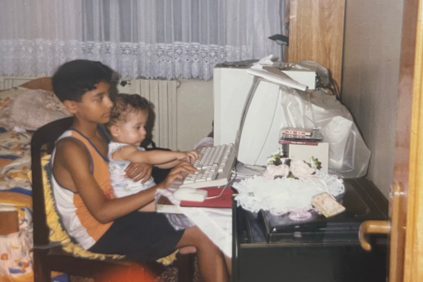 Çocuk yaştaki Okan Özalan aile bilgisayarının başında, bebek kardeşi Atakan'ı kucağında tutuyor; bebek, tüplü monitörlü bej bir bilgisayarın klavyesine uzanıyor.